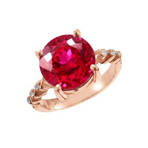 Può includere: Un anello in oro rosa con una grande gemma rossa brillante e rotonda. La fascia presenta gemme più piccole e trasparenti e un motivo strutturato. L'anello è ambientato su uno sfondo bianco.