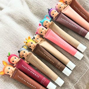 以下が含まれることがあります： 様々な色のSummer Fridays Butter Balmリップバームの詰め合わせ。各チューブには、気まぐれなフィギュアが飾られています。チューブは、ブランドのロゴと製品の詳細を示す、テクスチャ加工された表面に配置されています。