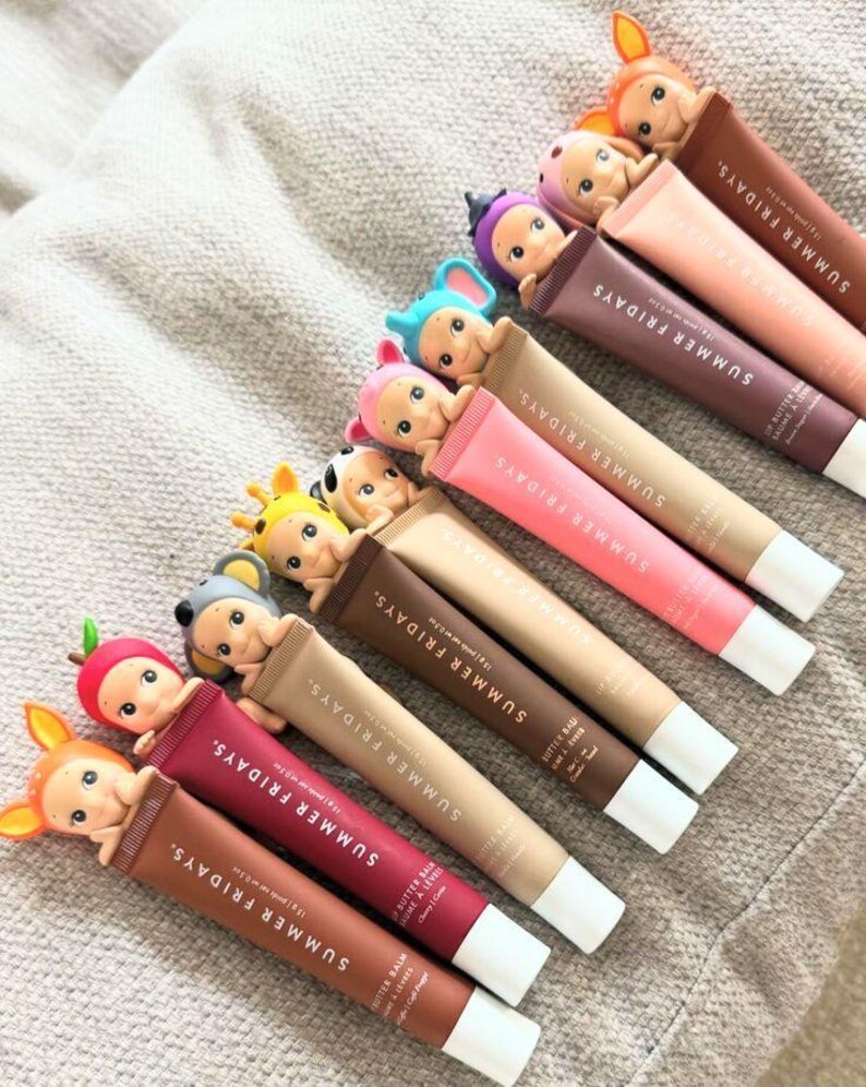 以下が含まれることがあります： 様々な色のSummer Fridays Butter Balmリップバームの詰め合わせ。各チューブには、気まぐれなフィギュアが飾られています。チューブは、ブランドのロゴと製品の詳細を示す、テクスチャ加工された表面に配置されています。