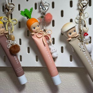 以下が含まれることがあります： リップバターのチューブから出てくる小さなフィギュアが特徴の、3つの装飾的なキーホルダー。フィギュアはキリン、ニンジン、キノコです。各キーホルダーは異なる色のチューブで、チャームと金属製の留め金で飾られています。