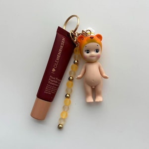 以下が含まれることがあります： オレンジ色のクマの帽子をかぶった小さな人形、濃い赤色のリップトリートメントチューブ、ビーズチャームが付いたキーホルダー。 リップトリートメントには「OLEHENRIKSEN」と「Pout Preserve Peptide Lip Treatment」と書かれています。 キーホルダーにはゴールドリングが付いています。