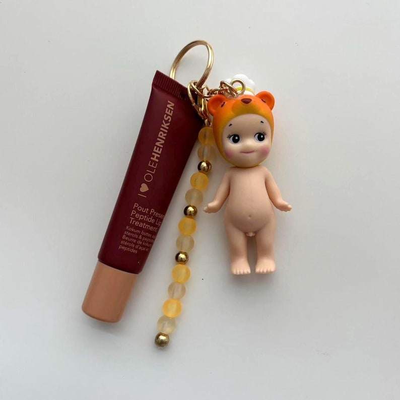 以下が含まれることがあります： オレンジ色のクマの帽子をかぶった小さな人形、濃い赤色のリップトリートメントチューブ、ビーズチャームが付いたキーホルダー。 リップトリートメントには「OLEHENRIKSEN」と「Pout Preserve Peptide Lip Treatment」と書かれています。 キーホルダーにはゴールドリングが付いています。