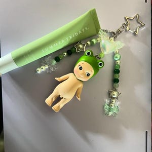 以下が含まれることがあります： サマーフライデーズのライトグリーンのチューブ製品と、カエルの帽子をかぶった小さな人形、グリーンのビーズ、星型のチャームが付いたチャーム。チャームには、透明な蝶と星型のキーホルダーも含まれています。