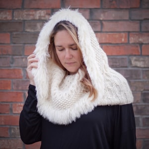 May include: A white knitted cowl with a faux fur trim. The cowl has a hood and is being worn by a person with blonde hair.