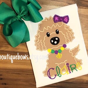 May include: A white t-shirt with a brown dog wearing a purple and yellow beaded necklace and a purple bow. The dog's name, "Claire", is embroidered in purple and yellow on the shirt. A green bow is also pictured.
