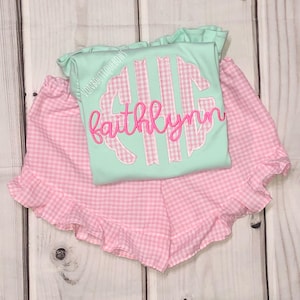 May include: A pink and white gingham ruffle shorts set with a light blue shirt featuring a monogram with the name "faithlynn" in pink.