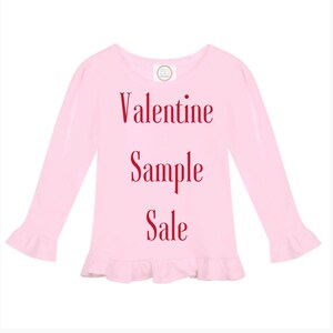 May include: A light pink long-sleeved shirt with a round neckline and ruffled cuffs and hem. The words "Valentine Sample Sale" are printed in red script across the front of the shirt.