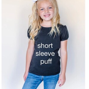 May include: A young girl wearing a black short-sleeved puff t-shirt with the words "short sleeve puff" printed on the front. She is also wearing blue jeans and a white bow in her hair.