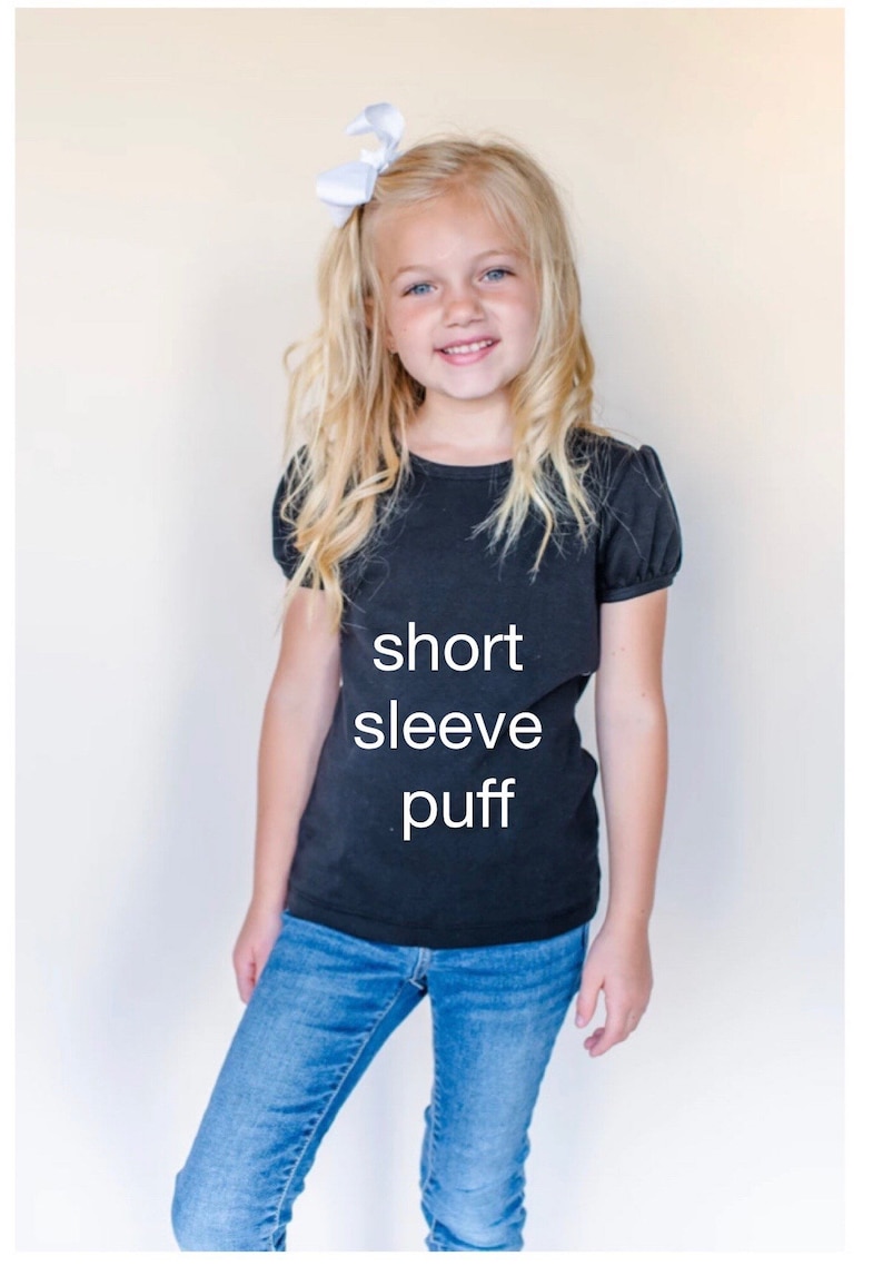 May include: A young girl wearing a black short-sleeved puff t-shirt with the words "short sleeve puff" printed on the front. She is also wearing blue jeans and a white bow in her hair.