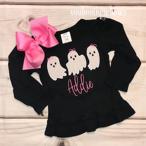 May include: Black long-sleeved shirt with a ruffle hem, featuring three embroidered ghosts with pink bows and the name "Addie" in pink lettering.