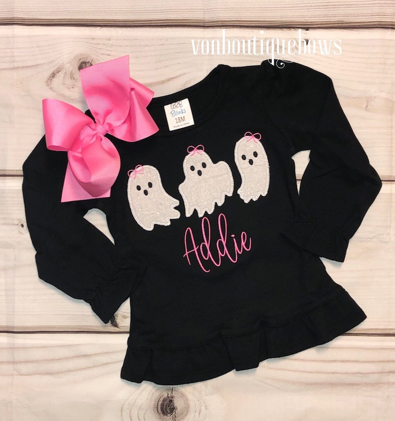 May include: Black long-sleeved shirt with a ruffle hem, featuring three embroidered ghosts with pink bows and the name "Addie" in pink lettering.