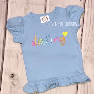 May include: A light blue short-sleeved shirt with a ruffled hem. The shirt has the name "sterling" embroidered on the front in rainbow colors with a yellow heart.