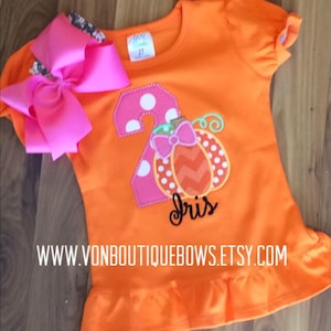 May include: Orange short-sleeved shirt with a pink number two and a pumpkin design. The shirt has a ruffle at the bottom and says "Iris" on the front. The shirt is for a child.