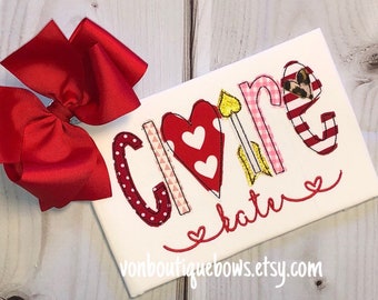 May include: A white t-shirt with the word "Love" spelled out in colorful letters with hearts and arrows. The letters are decorated with different patterns, including polka dots, stripes, and gingham. The name "kate" is written in cursive below the word "Love".