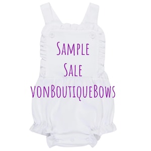 May include: White baby romper with ruffle trim, shoulder straps, and snap closure. The text "SAMPLE SALE VONBOUTIQUEBOWS" is printed in purple across the front. Buttons are visible on the front.
