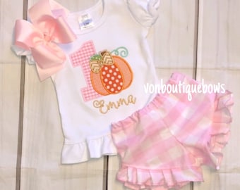 May include: A white short-sleeved shirt with a pink and white gingham number one and a pumpkin with orange, white, and green accents. The shirt has a ruffle at the bottom and the name "Emma" is embroidered below the pumpkin. The shirt is paired with pink and white gingham shorts with a ruffle at the bottom.