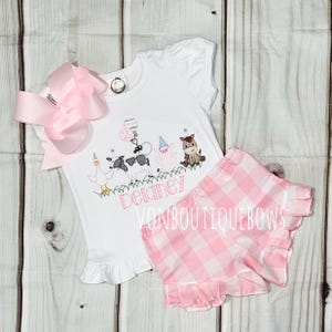 May include: White short-sleeved shirt and pink gingham shorts set. The shirt features an embroidered farm animal design with the name "Delaney" and a large pink bow. The shorts have a ruffled hem.