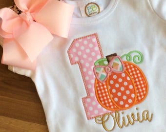 Può includere: Body bianco con un numero uno a pois rosa e bianchi e una zucca con pois arancioni e bianchi e un motivo a quadri. La zucca ha uno stelo verde e un fiocco verde e rosa. Il nome Olivia è ricamato in oro sotto la zucca. Il body ha un volant bianco in fondo.