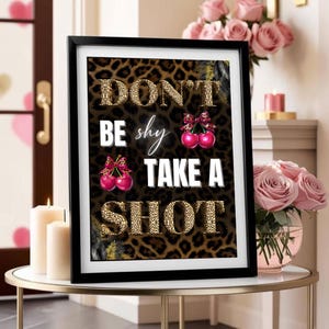May include: A framed art print with the text "DON'T BE shy TAKE A SHOT" in a leopard print and white font. The image includes illustrations of cherries with bows. The frame is black, and the print is displayed on a small table.