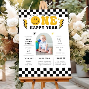 May include: A wooden easel displays a personalized first birthday sign. The sign features a photo of a baby, the text "ONE HAPPY YEAR", and details like weight (22 pounds) and height (31 inches). The sign has a black and white checkered border.