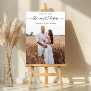May include: A wooden easel displays a welcome sign with the text "WELCOME TO the night before ISABELLA & THEODORE." The sign features a photo of a couple in a wheat field. A vase of dried pampas grass sits to the left.