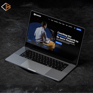 May include: A silver laptop showing a website with the text "INBUSINESS". The screen displays a man at a podium with the words "Coaching for Business Growth to be more Positive". The laptop is on a dark surface.