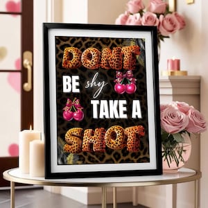 以下が含まれることがあります： ヒョウ柄のフォントで「DON'T BE shy TAKE A SHOT」という言葉が書かれた額入りのプリント。背景はヒョウ柄模様です。画像にはリボンが付いたサクランボのイラストが含まれています。