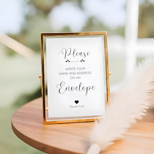 May include: A gold-framed sign with the words "Please write your name and address on an envelope" in elegant script. A small black heart and the words "Thank You!" are at the bottom. The sign is on a wooden table.