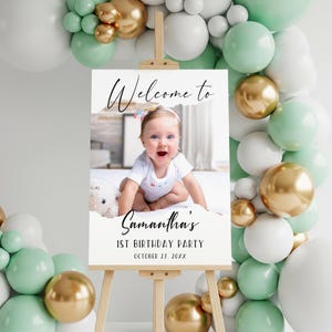 May include: A white sign on a wooden easel with the text "Welcome to Samantha's 1st Birthday Party October 27, 20XX". The sign features a photo of a baby girl. The background includes balloons in white, gold, and mint green.
