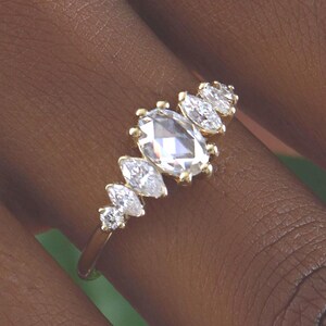 May include: A gold ring featuring an oval-cut center stone and several smaller, marquise-cut stones. The ring is set in a gold band and is displayed on a person's finger. The ring's design is elegant and eye-catching.