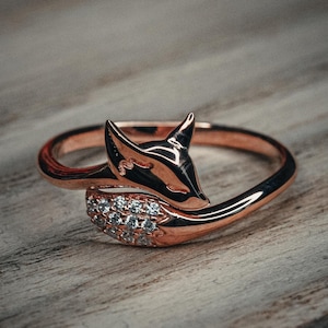 May include: A rose gold ring featuring a fox design. The fox head is embellished with small, clear, sparkling stones. The ring is set against a wooden surface.