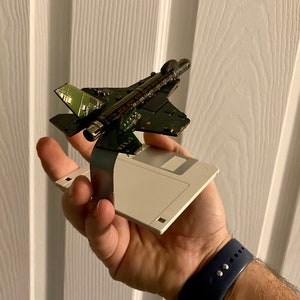 May include: A small model airplane made from circuit boards is held in a hand. The airplane is perched on a floppy disk.
