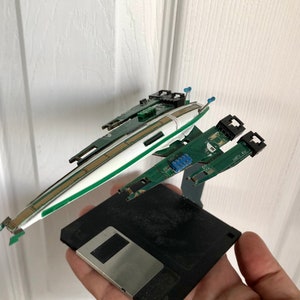 May include: A model spaceship made from recycled computer parts, including circuit boards and a floppy disk. The spaceship is green, white, and black.