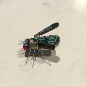 May include: A small, insect-like sculpture made from recycled electronic components. The sculpture is green, brown, and silver with red accents. The wings are made from circuit boards.