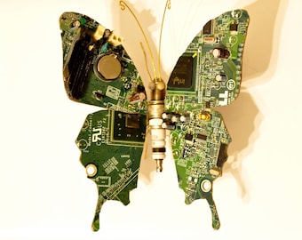 Puede incluir: Una escultura de mariposa de placa de circuito verde con una antena de alambre dorado y una bujía en el centro.