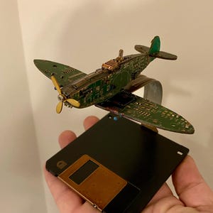 May include: A model airplane made from recycled circuit boards and other materials. The plane is green, brown, and gold and is sitting on a black floppy disk.