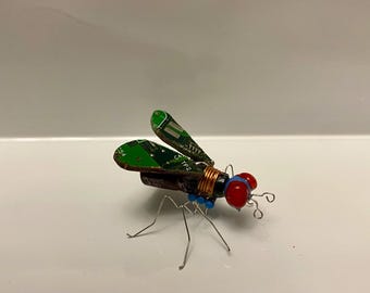 Puede incluir: Una pequeña escultura de insecto verde, azul y rojo hecha de placas de circuito recicladas y alambre. El insecto tiene dos alas grandes, un cuerpo pequeño y dos ojos rojos.