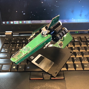 May include: A green circuit board spaceship model with black capacitors and a floppy disk on a black laptop keyboard.