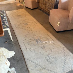 May include: A long, rectangular rug with a faded, vintage appearance. The rug features a central geometric pattern in shades of beige, grey, and blue. The text "ZS CarpetsIN" is visible on the rug. The rug is placed on a concrete floor.