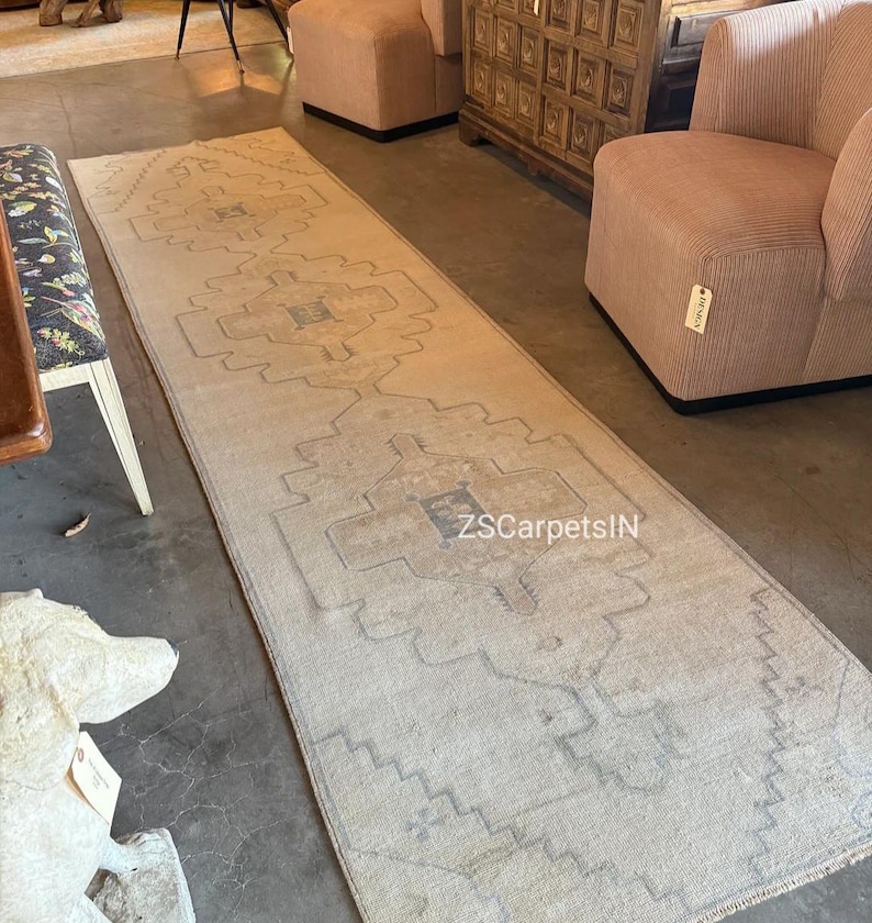 May include: A long, rectangular rug with a faded, vintage appearance. The rug features a central geometric pattern in shades of beige, grey, and blue. The text "ZS CarpetsIN" is visible on the rug. The rug is placed on a concrete floor.