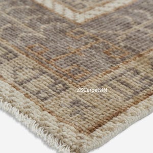 May include: Close-up of a beige and brown patterned rug with a geometric design. The rug has a fringe along the edge and the text "ZSCarpetsIN" is visible. The rug is made of a woven material.