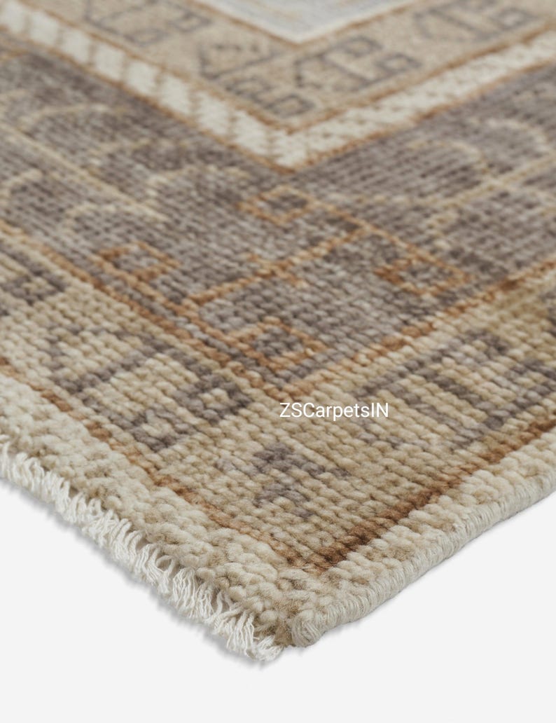May include: Close-up of a beige and brown patterned rug with a geometric design. The rug has a fringe along the edge and the text "ZSCarpetsIN" is visible. The rug is made of a woven material.