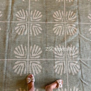 Könnte beinhalten: Ein salbeigrüner Teppich mit einem sich wiederholenden Gittermuster und cremefarbenen muschelartigen Motiven in jedem Quadrat. Der Teppich hat eine gewebte Textur und enthält den Text ZSCarpetsIN, der in Weiß auf die Stoffoberfläche gedruckt ist.