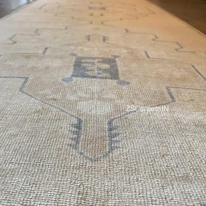 May include: Beige and grey patterned rug. The rug features a central design with geometric shapes and a border. It is made of a woven material with a textured surface. The rug is rectangular and appears long and narrow.