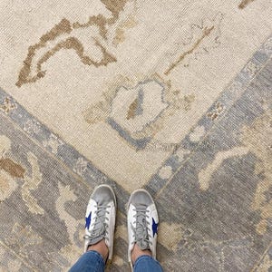 Könnte beinhalten: Draufsicht auf einen beige-grauen gemusterten Teppich mit Bordüre. Der Teppich hat einen Used-Look mit floralen und geometrischen Designs. Eine Person trägt blaue Jeans und weiße Turnschuhe mit blauen Sternakzenten.