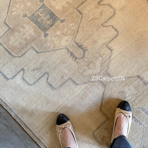 May include: Beige and gray patterned rug with a geometric design. The rug has a central diamond shape with floral elements. The text "ZSCarpetsIN" is visible. A person is wearing beige flats with black toes and blue jeans.