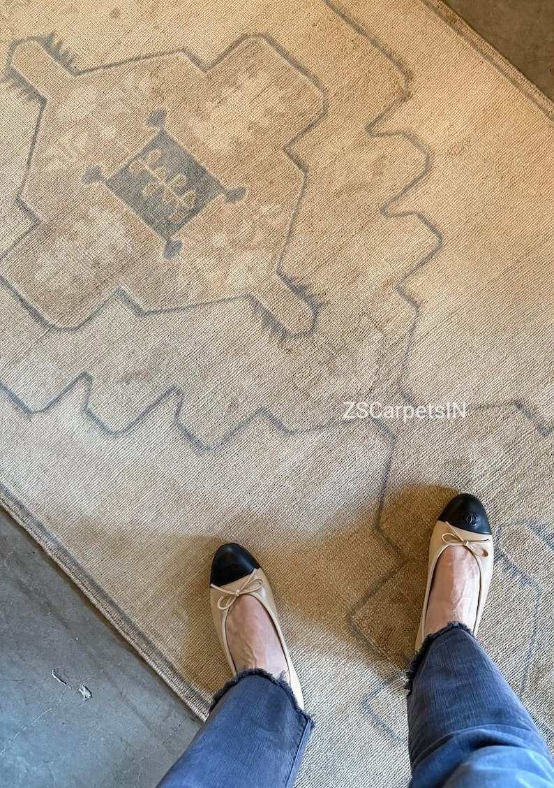 May include: A beige and grey patterned rug with a geometric design. The rug features a central diamond shape with floral elements. The text "ZSCarpetsIN" is visible. A person is wearing beige flats with black toes and blue jeans.