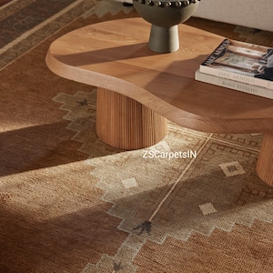 May include: A light brown, oval-shaped wooden coffee table with a fluted base. A dark green bowl sits on top, along with two books. A patterned brown and tan rug is visible in the foreground. The text "ZSCarpetsIN" is visible on the rug.
