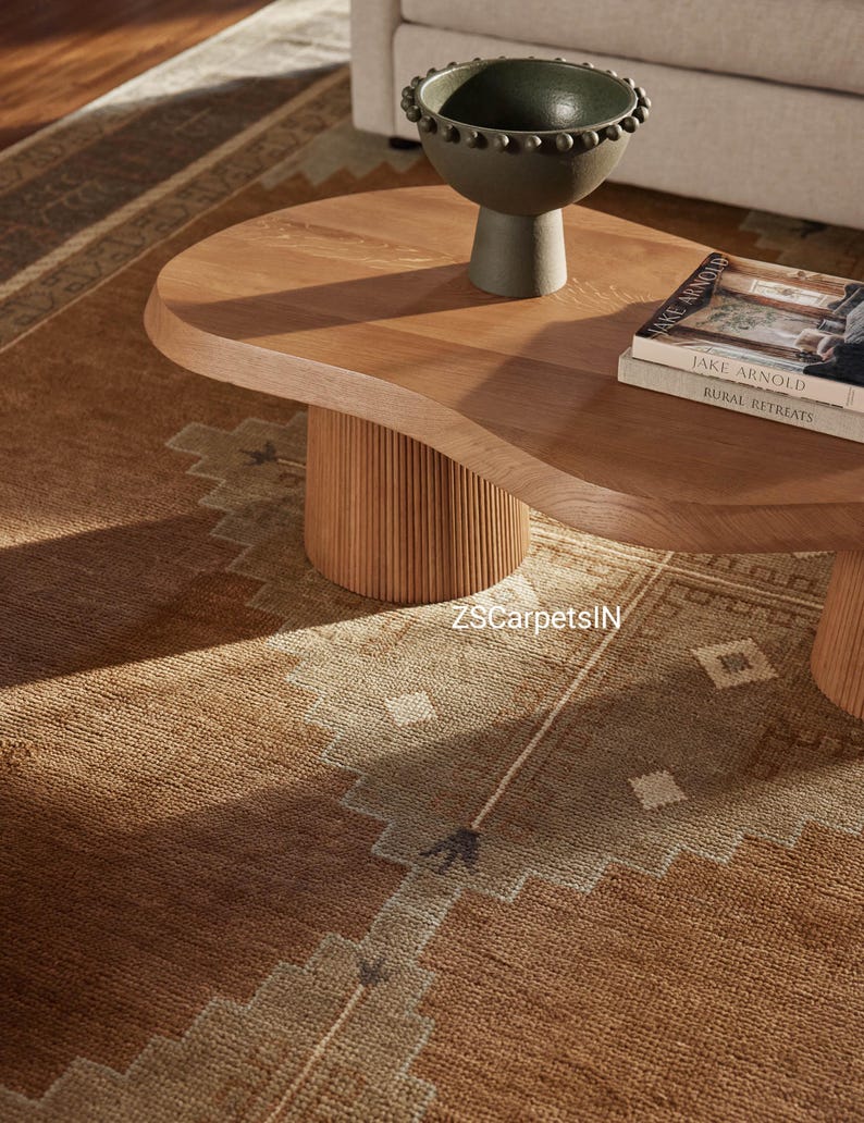 May include: A light brown, oval-shaped wooden coffee table with a fluted base. A dark green bowl sits on top, along with two books. A patterned brown and tan rug is visible in the foreground. The text "ZSCarpetsIN" is visible on the rug.