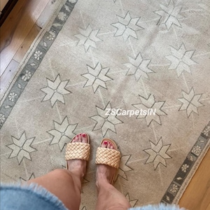May include: A beige rug with a repeating star pattern and a gray border. The rug is on a wooden floor. Woven sandals with a woven design are visible.